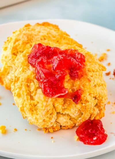 A close up picture of two buttermilk biscuits on a white plate, one with strawberry jam on it.