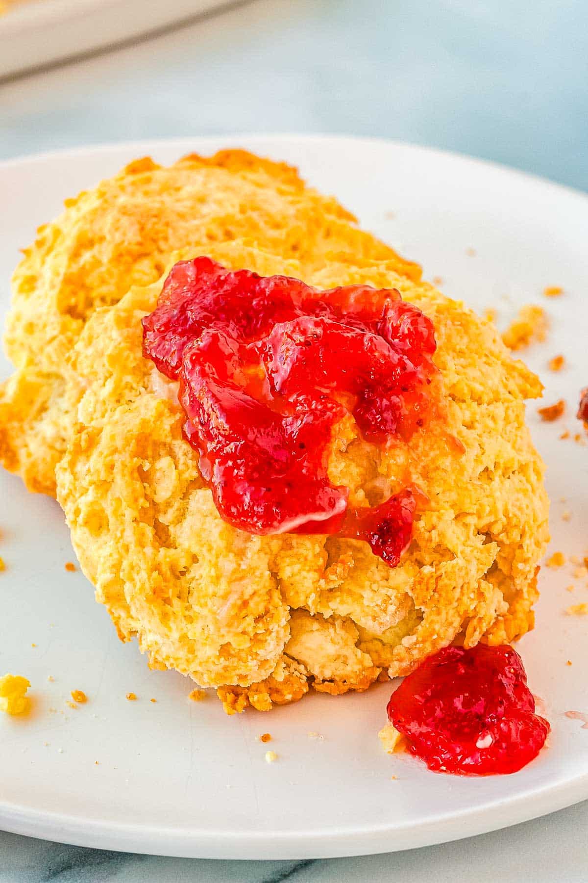 A close up picture of two buttermilk biscuits on a white plate, one with strawberry jam on it. 