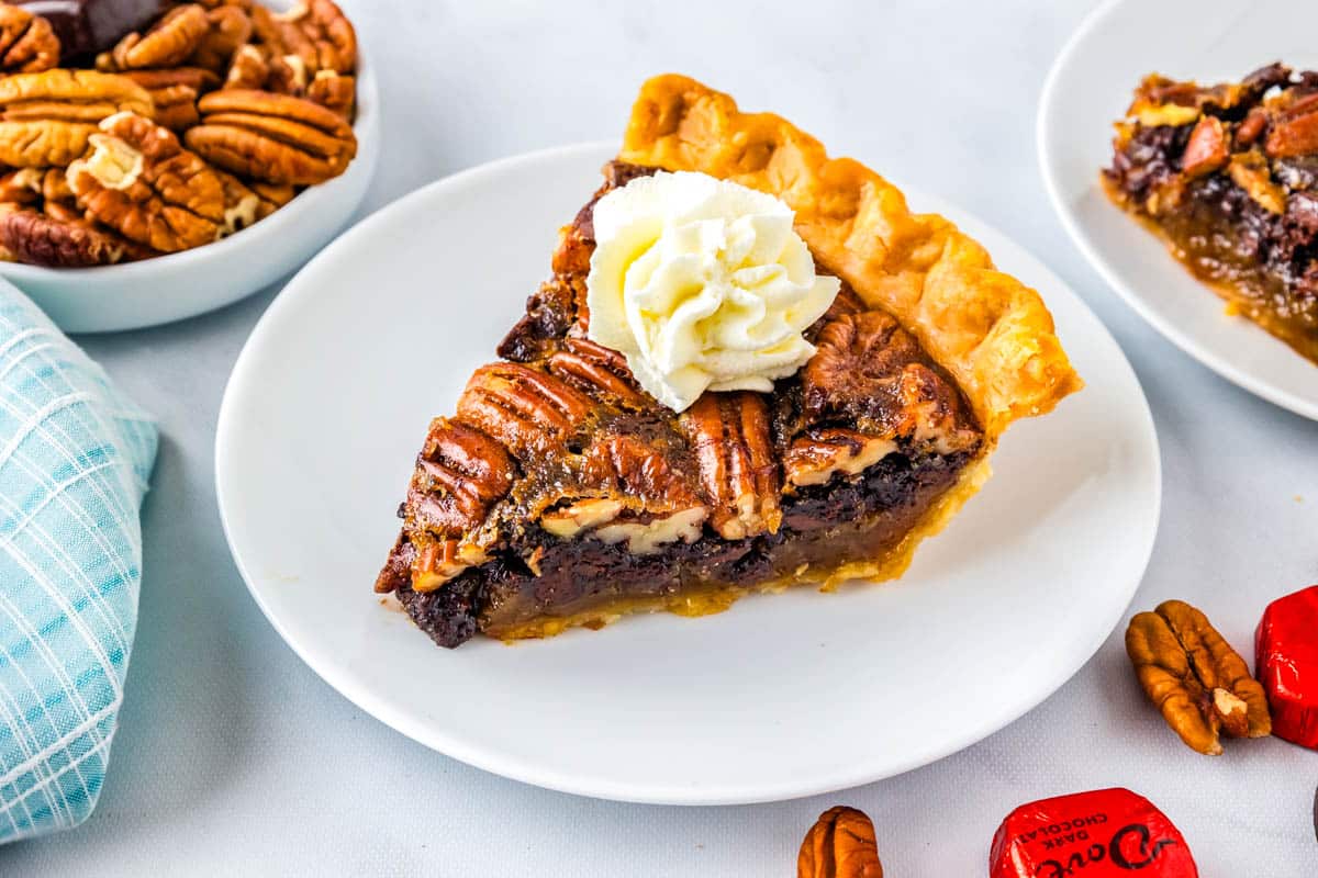 A slice of dark chocolate pecan pie on a white plate with whipped cream.