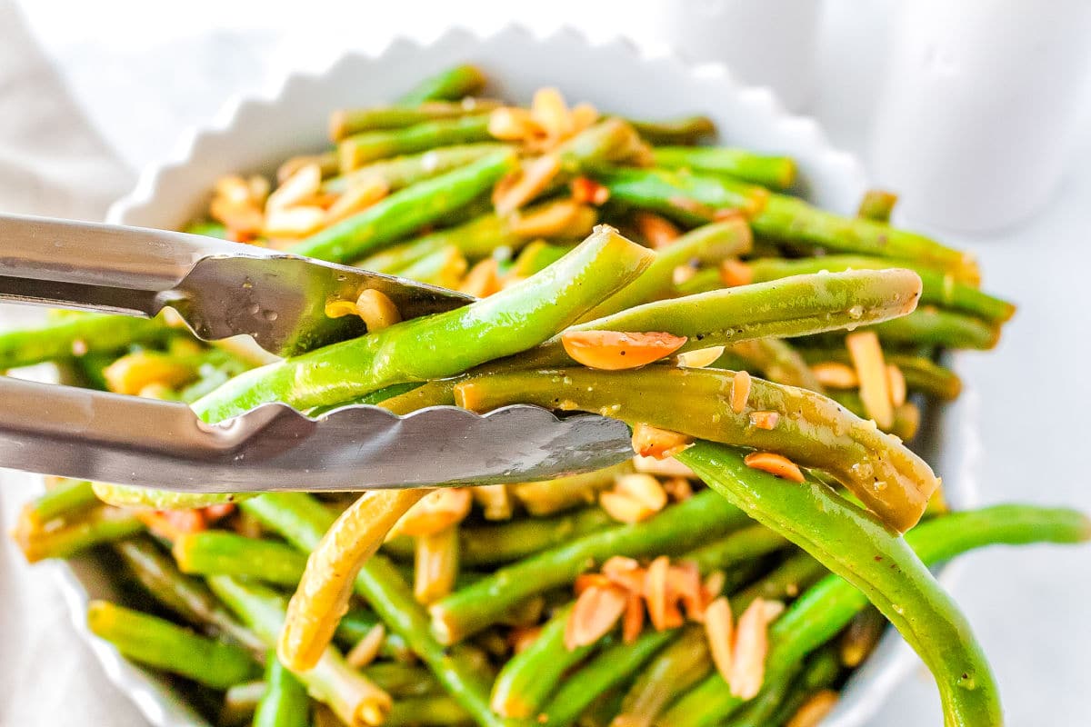 tongs grabbing a scoop of green beans from a white serving bowl
