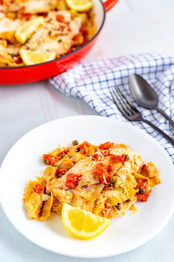 Tilapia on a white plate with tomatoes, artichoke hearts, and lemon wedge. 