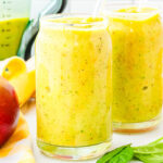 two glasses of mango smoothie with a blender, banana, and mango in the background