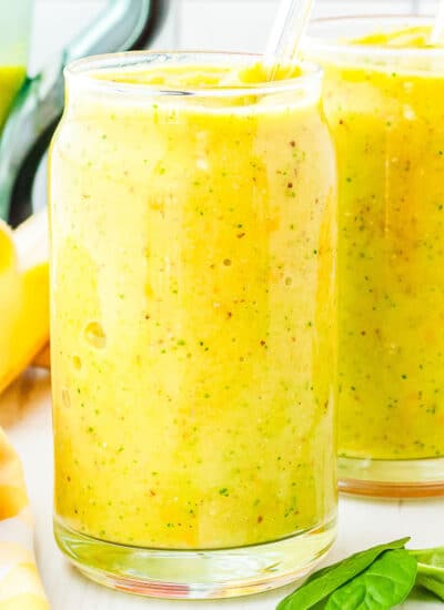 two glasses of mango smoothie with a blender, banana, and mango in the background