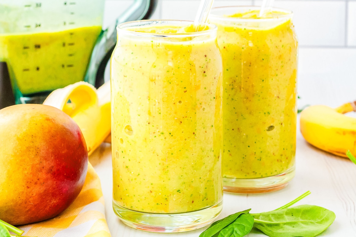 two glasses of mango smoothie with a blender, banana, and mango in the background