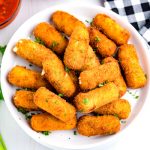 Fried Mozzarella Cheese Sticks on a white plate with dipping sauce in the background.