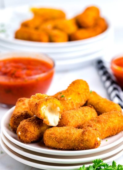 A pile of Mozzarella Cheese Sticks on a white plate with marinara sauce in the background.