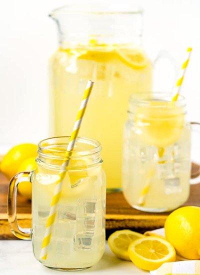 Lemonade that is homemade in mason jar cups and a large pitcher and lemons in the background.
