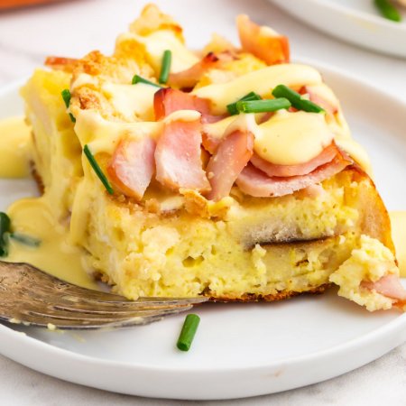 A close up picture of a slice of eggs benedict casserole on a white plate with a fork.