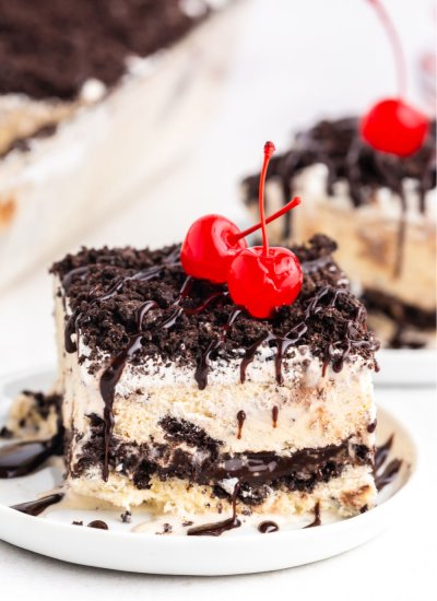 A slice of Oreo Ice Cream Cake on a white plate topped with hot fudge and cherries.
