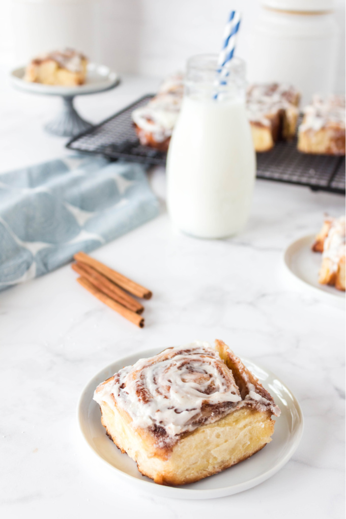 A cinnamon roll on a white plate with milk and the rest of the overnight cinnamon rolls in the background.