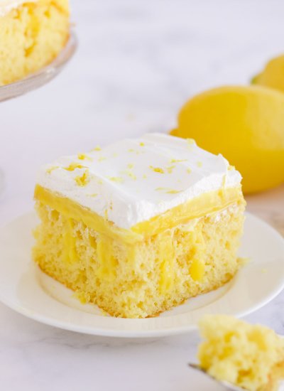 A piece of lemon cake on a plate. Lemons in the background. A piece of cake on a fork