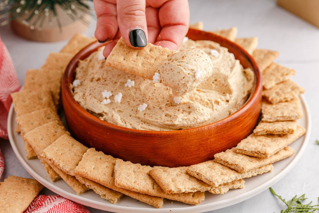 A picture of a hand scooping some of the Gingerbread Cheesecake Dip with a graham cracker.