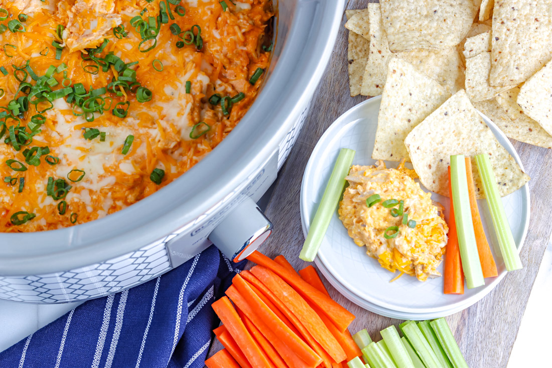 An overhead picture of the finished Crockpot Buffalo Chicken Dip in a slow cooker and on a plate with some chips and carrots.