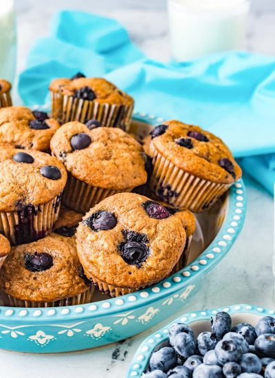 The finished Banana Blueberry Muffins in a blue serving platter with blueberries and milk nearby.