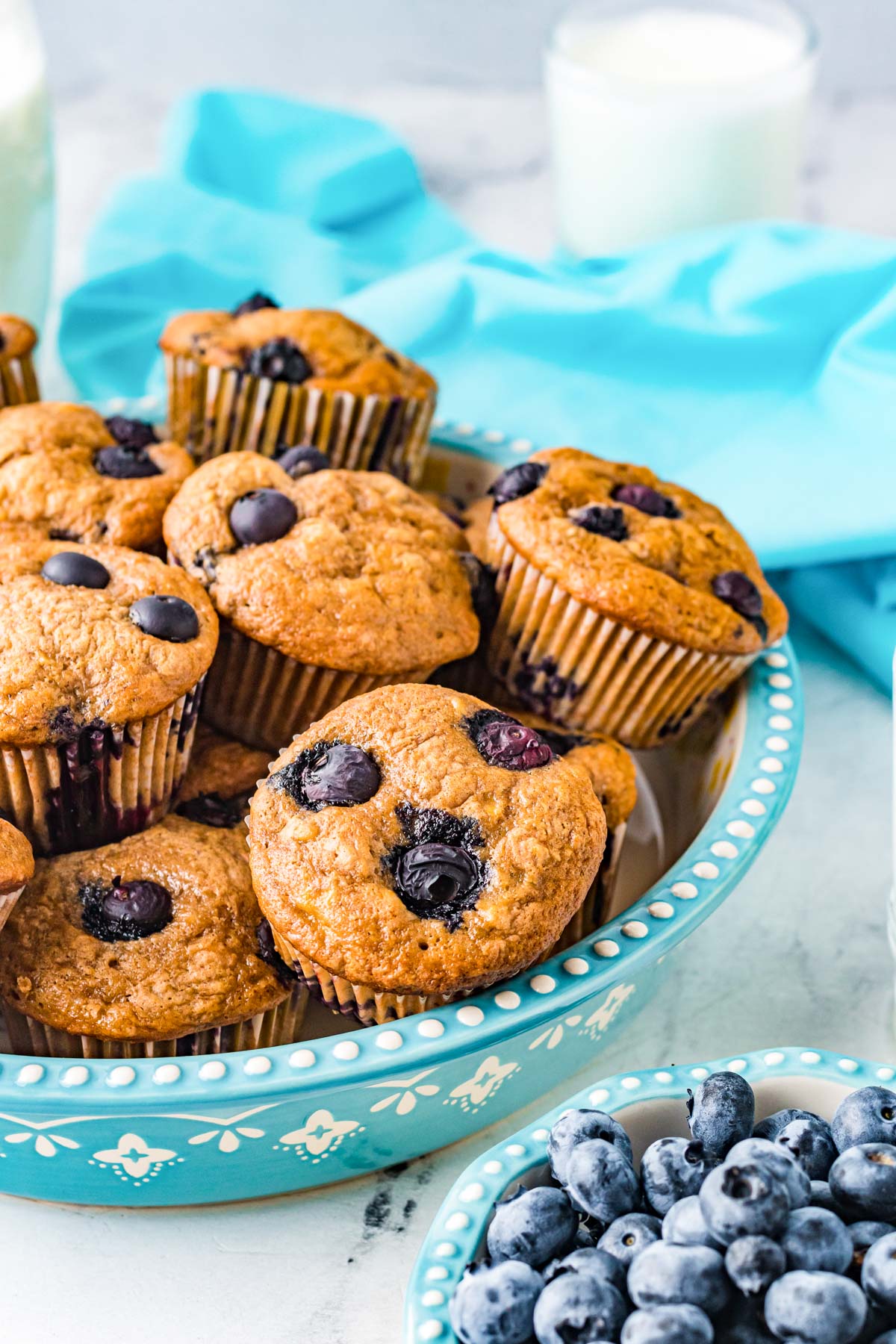 The finished Banana Blueberry Muffins in a blue serving platter with blueberries and milk nearby.