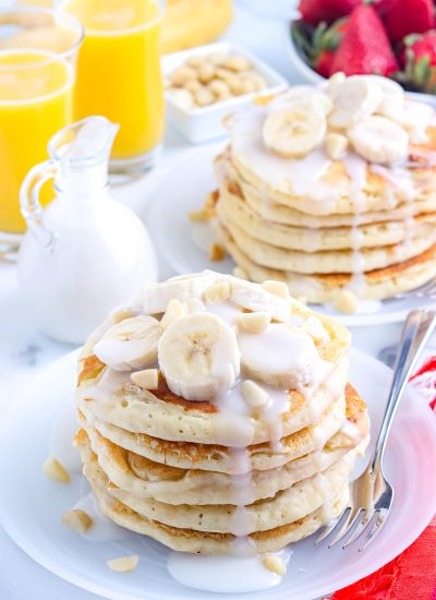 Two stacks of the finished Banana Pancakes on plates topped with sliced bananas, macadamia nuts, and coconut syrup.