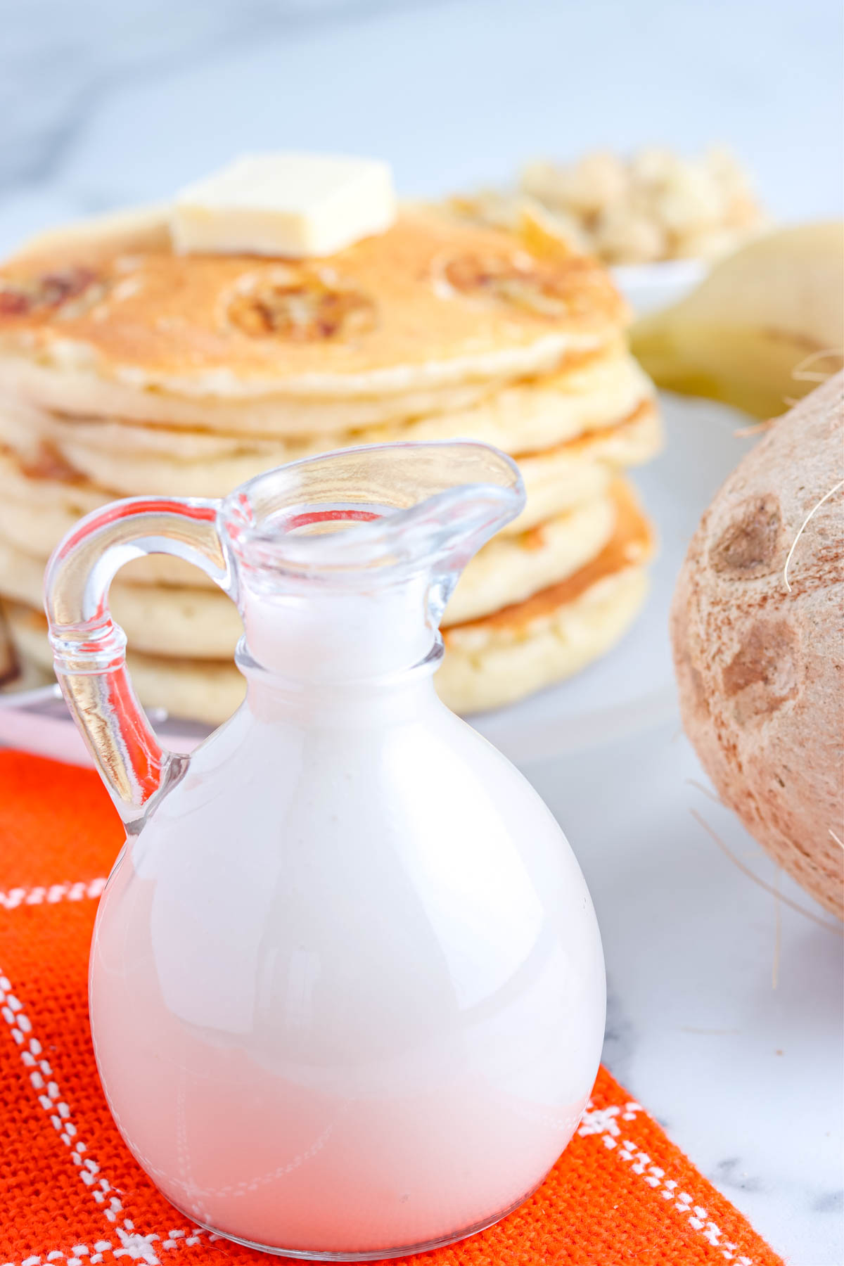 A small glass pitcher full of coconut syrup with banana pancakes in the background. 