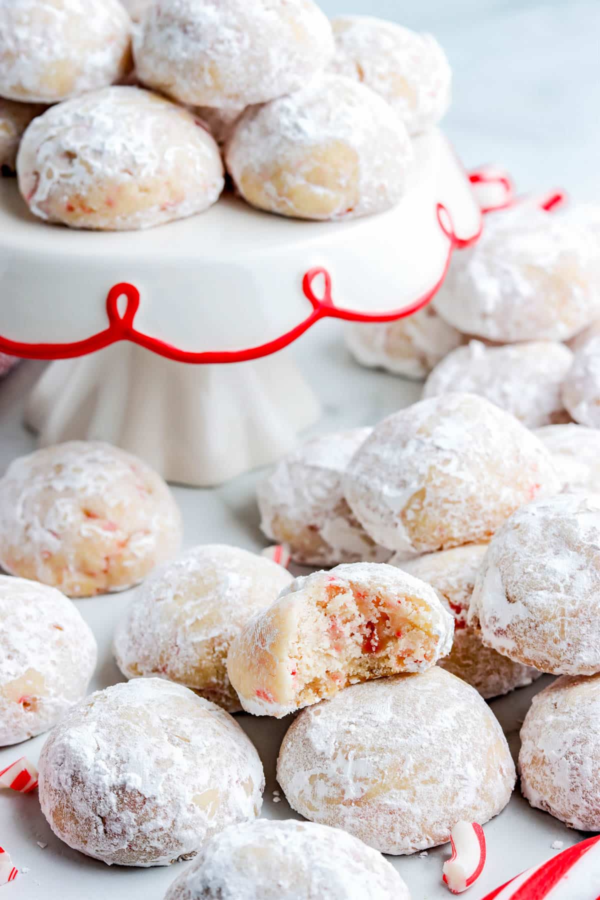 A pile of Peppermint Snowball Cookies on a counter and on a cake stand.