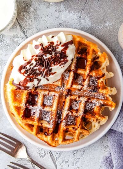 overhead picture of waffles on a plate that are garnished with chocolate sauce, whipped cream, and chocolate sprinkles.