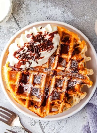 overhead picture of waffles on a plate that are garnished with chocolate sauce, whipped cream, and chocolate sprinkles.