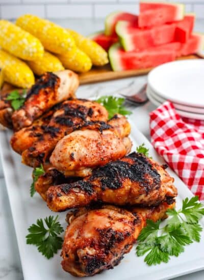 Chicken drumsticks on a white platter with corn and watermelon in the background.