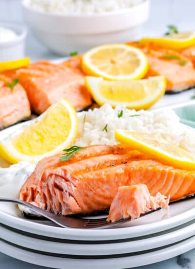 a fork taking a bite of salmon from a plate with a salmon fillet on it and a platter with more salmon on it in the background