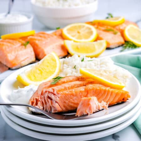 a fork taking a bite of salmon from a plate with a salmon fillet on it and a platter with more salmon on it in the background