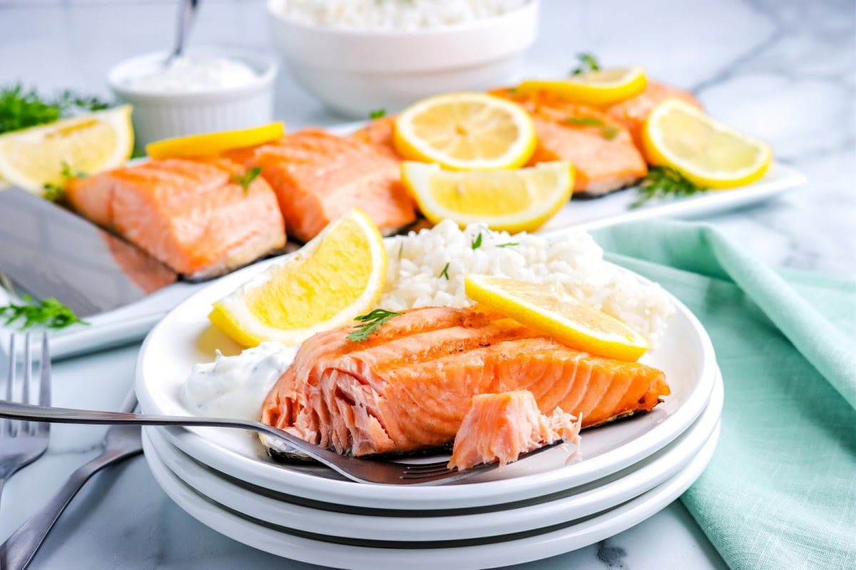 a fork taking a bite of salmon from a plate with a salmon fillet on it and a platter with more salmon on it in the background