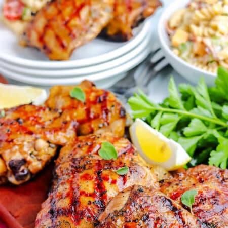 a platter of grilled chicken thighs with some pasta salad and a plate of chicken thighs in the background