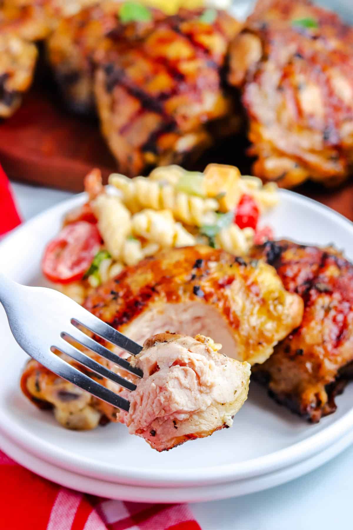 a fork taking a bite of chicken from the grilled chicken thigh on the plate with pasta salad in the background