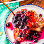 a ladle of blueberry syrup being poured over a plate of French toast