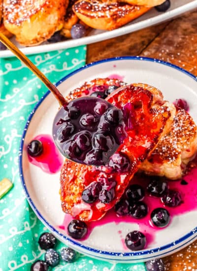 a ladle of blueberry syrup being poured over a plate of French toast