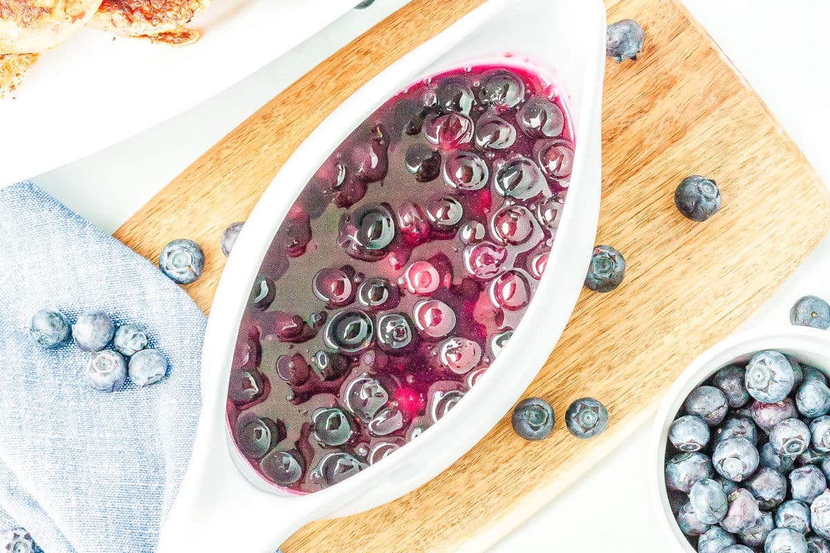 a gravy boat full of blueberry syrup on a cutting board with blueberries on the side