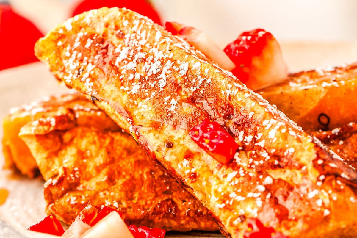 a close up of a French toast stick with others and strawberry slices in the background on a plate