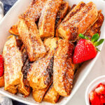 a plate of french toast sticks with strawberry and orange slices in the background