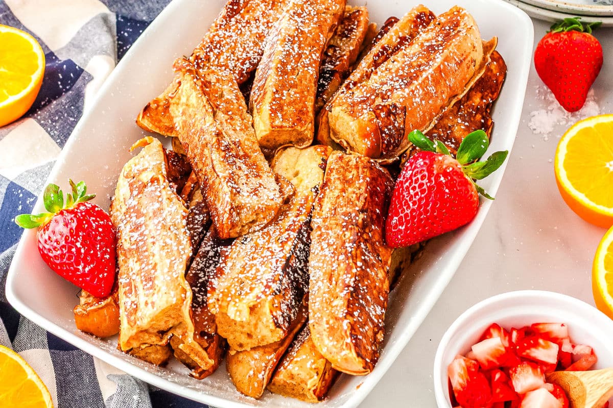 a plate of french toast sticks with strawberry and orange slices in the background
