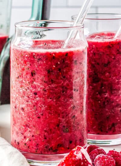 two glasses with smoothies in them with a blender, fruit, and kale in the background
