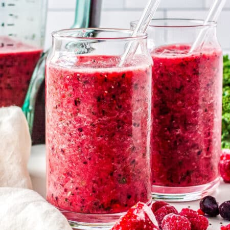 two glasses with smoothies in them with a blender, fruit, and kale in the background