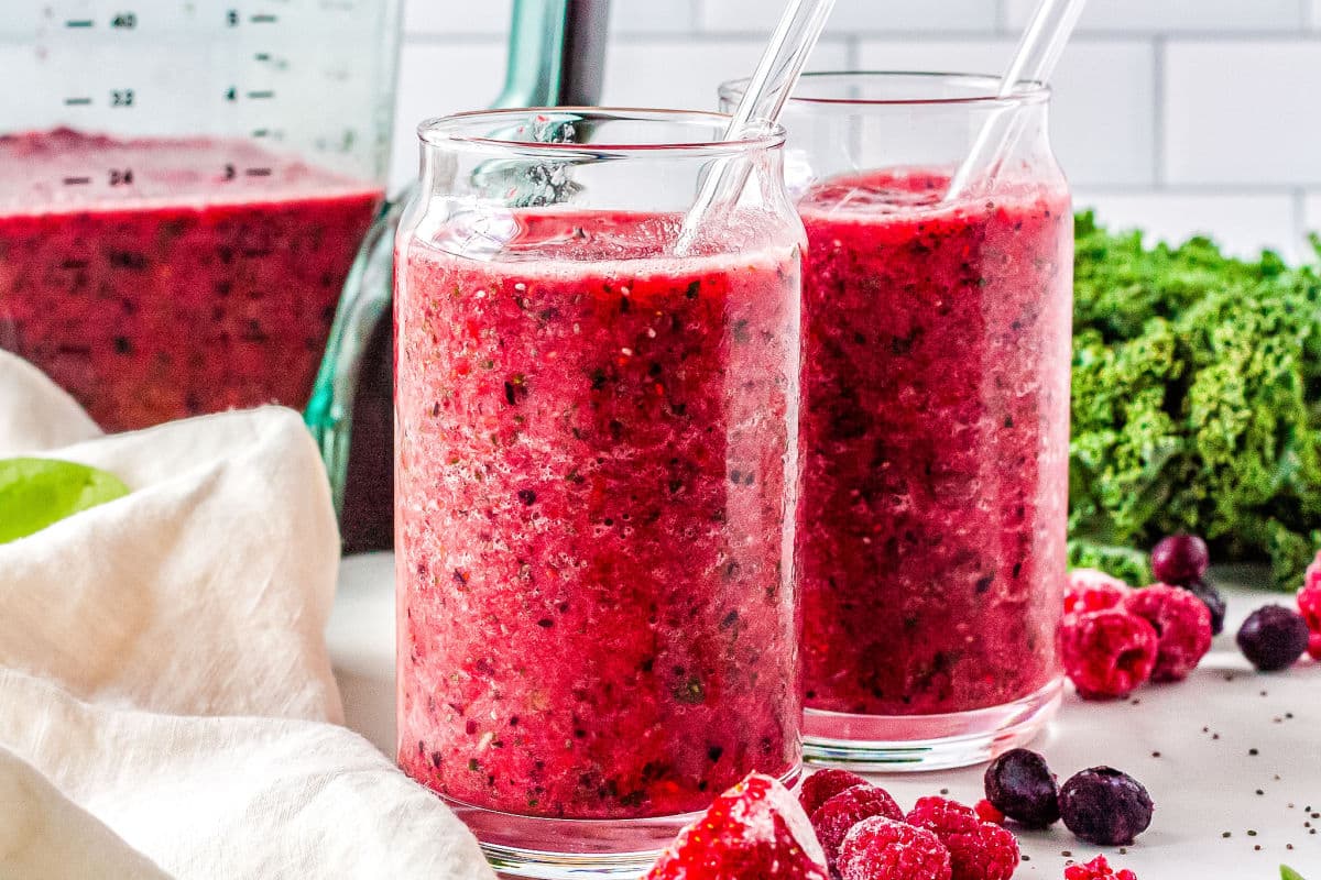 two glasses with smoothies in them with a blender, fruit, and kale in the background
