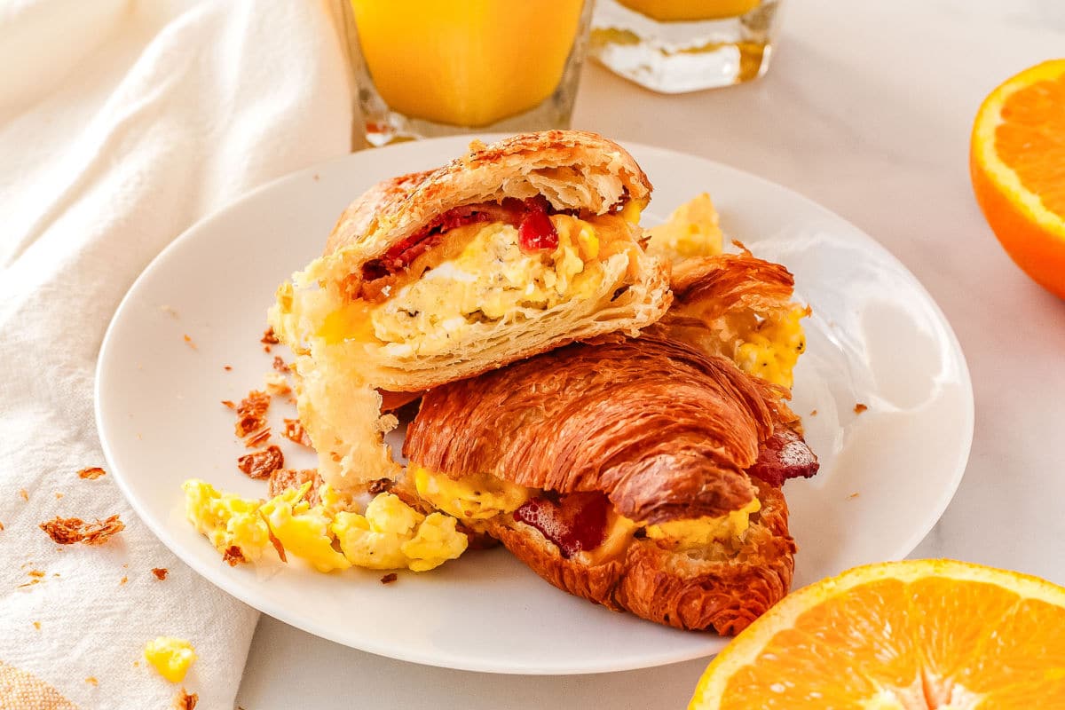 a baked breakfast crossiant sandwich on a plate with some sliced oranges in the background