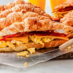 a close up of a non-baked breakfast sandiwch being picked up by a spatula, with more croissant sandwiches in the background