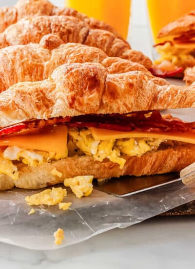 a close up of a non-baked breakfast sandiwch being picked up by a spatula, with more croissant sandwiches in the background