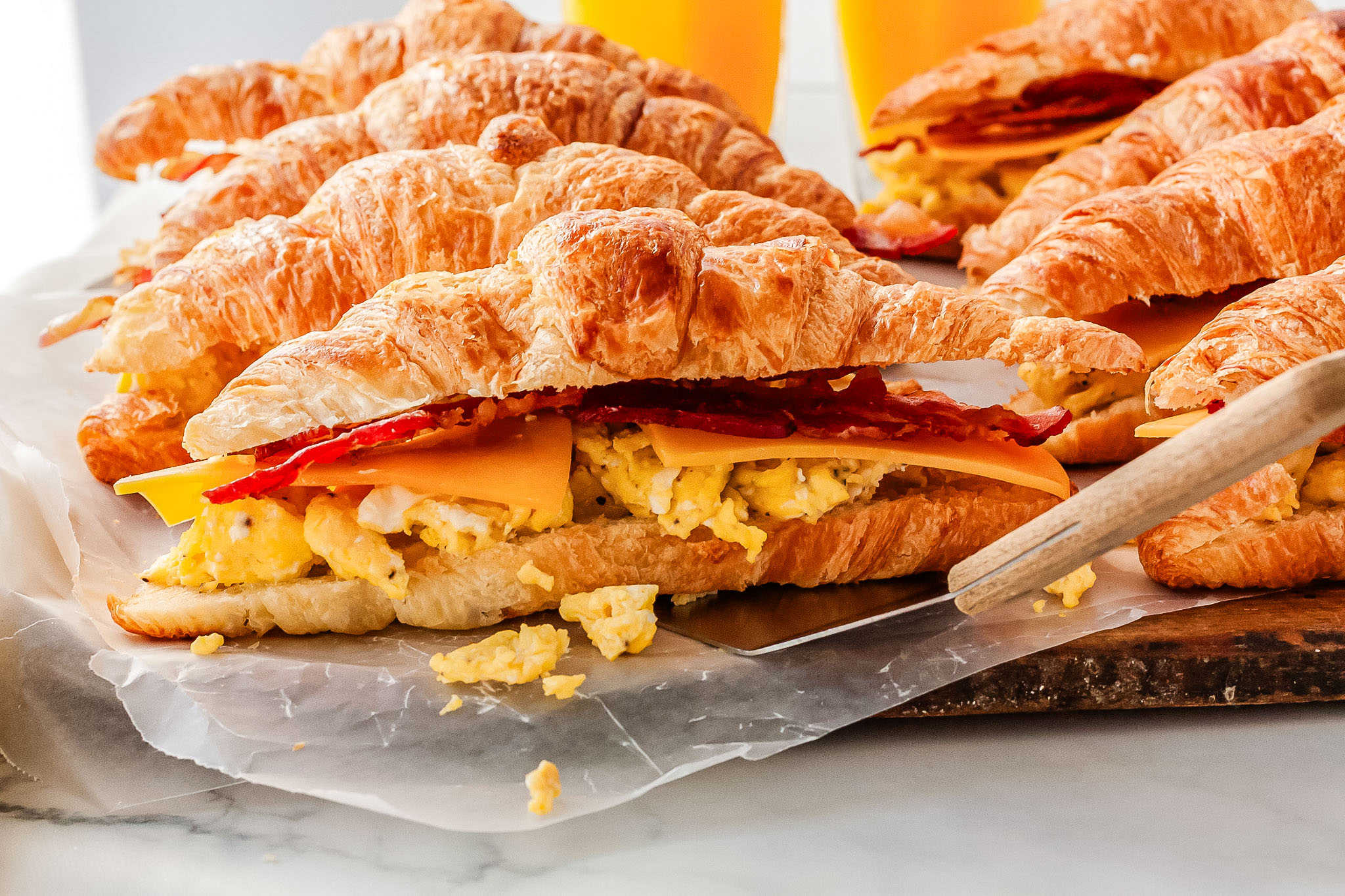 a close up of a non-baked breakfast sandiwch being picked up by a spatula, with more croissant sandwiches in the background